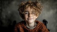 Young boy with curly blonde hair wearing orange knit sweater smiling warmly at camera in cozy indoor setting for lifestyle and family content.