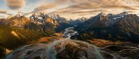 Aerial view of winding river flowing through alpine valley surrounded by snow-capped mountains and forested slopes during golden hour sunset landscape photography.