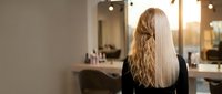 Young blonde woman with long wavy hair sitting in modern hair salon chair facing mirror with warm lighting and professional styling stations.