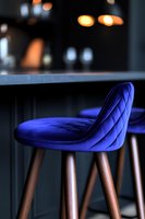 Modern bar stool with quilted royal blue velvet upholstery and wooden legs against dark background with soft bokeh lights creating luxury atmosphere.