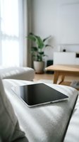 Modern tablet device resting on soft gray sofa in bright minimalist living room with natural light streaming through window and green plant in background.