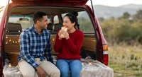 Happy mixed-race couple enjoying romantic picnic date in car trunk during outdoor adventure trip in scenic countryside landscape at golden hour sunset.