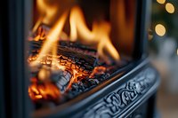 Burning logs in ornate cast iron fireplace with decorative details, glowing orange flames and embers creating cozy atmosphere on dark background.