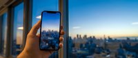 Young adult hand holding smartphone displaying cityscape view from high-rise window, capturing urban skyline at dusk for travel, technology, and lifestyle content.
