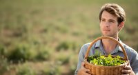 Young Caucasian male farmer holding wicker basket of fresh leafy greens in agricultural field during harvest season.