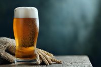 Fresh draft beer in glass with thick foam head and wheat ears on rustic wooden table against dark background, cold bubbling lager beverage.