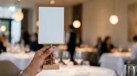 Cropped hand holding blank white card on wooden stand at formal dinner event with guests and table settings in blurred background for messaging.