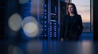 Professional businesswoman standing confidently in modern data center with server racks and bokeh lights creating atmospheric technology environment.