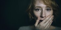 Young Caucasian woman with fearful expression covering mouth with hands against dark background, portraying anxiety, fear, or silence.