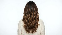 Back view of woman with long wavy brown hair showing natural highlights and layers, wearing cream sweater against white background for hair styling concepts.