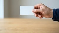 Male hand holding blank white business card over wooden table surface with blurred background, suitable for corporate branding and professional mockup designs.