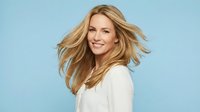Young blonde Caucasian woman with flowing hair in white blazer smiling confidently against light blue background for professional business portrait.