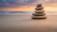 Zen stone stack on sandy beach at sunset with soft ocean waves and colorful sky creating peaceful meditation atmosphere for wellness and mindfulness practices.
