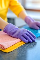 Hands in purple rubber gloves organizing colorful microfiber cleaning cloths on granite countertop, close-up view showing texture and detail of housekeeping supplies.