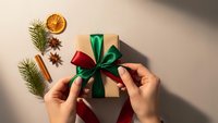 Hands tying festive ribbon on kraft paper gift box with pine branches, dried orange slice, cinnamon stick and star anise for holiday wrapping.