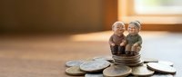 Elderly couple figurines sitting on stack of coins representing retirement savings and financial planning for senior years in warm sunlight.