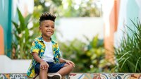 Young Black boy with curly hair wearing colorful tropical shirt sitting outdoors among plants, smiling at camera. Child portrait for parenting, family lifestyle, and summer activity content.