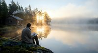 Man sitting on rocky shore watching sunrise over misty lake with cabin and forest reflection creating peaceful morning atmosphere for relaxation content.