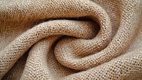 Natural linen fabric with twisted folds creating spiral texture pattern. Organic cotton or hemp textile in beige color showing detailed weave structure.