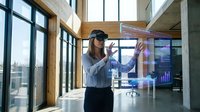 Young businesswoman using virtual reality headset interacting with holographic data displays in modern office environment with large windows and urban view.
