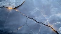 Dramatic cracked ice surface with glowing golden light shining through fissures against moody blue background creates abstract winter nature pattern.