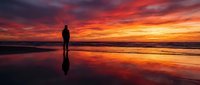 Silhouetted figure standing on wet beach at dramatic sunset with vibrant orange and red sky reflecting in water, suitable for inspirational and travel content.