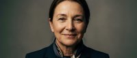 Middle-aged woman with dark hair, fair complexion, wearing dark blazer over striped shirt. Professional portrait against neutral gray background suitable for corporate, business, and editorial use.