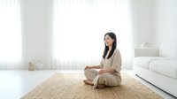 Young woman of East Asian descent sitting in lotus position on natural fiber mat in minimalist bright interior space, practicing meditation or yoga for wellness and mindfulness.