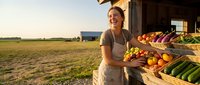 Young Caucasian woman farmer in apron holding fresh produce basket at rural farm stand with vegetables and fruits display during golden hour sunset.