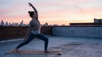 Young woman practicing yoga warrior pose on urban rooftop at sunset with city skyline background for wellness and fitness lifestyle content.