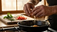 Professional chef hands cracking fresh egg into hot skillet pan for cooking breakfast in modern kitchen with herbs and tomatoes on wooden cutting board.