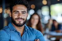 Young Hispanic man with beard wearing denim shirt smiling confidently with arms crossed in casual office or cafe setting with blurred background and bokeh lights.