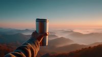 Hand holding travel mug against breathtaking mountain landscape at dawn, with fog-filled valleys and warm sunrise colors creating serene outdoor atmosphere.