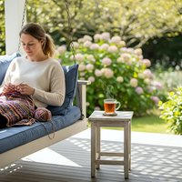 Young woman knitting on outdoor porch swing with tea cup, relaxing in sunny garden with blooming flowers, peaceful hobby and leisure lifestyle moment.