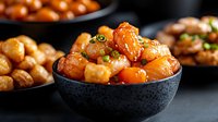 Sweet and sour chicken pieces glazed with honey sauce in black ceramic bowl, garnished with green onions against dark background, macro food photography.