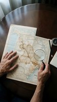 Mature adult male hands examining vintage geographical maps with magnifying glass on wooden table for exploration and research purposes.