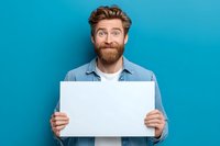Surprised man with red beard holding blank white poster against turquoise background, perfect for adding custom text or promotional message.