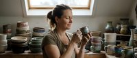 Caucasian female potter admires handmade ceramic mug in cozy studio surrounded by handcrafted bowls and vases on wooden shelves.