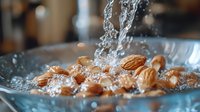 Fresh almonds being washed in clear water with dynamic splashes and droplets in glass bowl, macro food photography showing texture and detail of nuts.