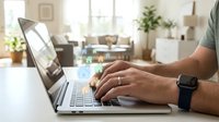 Man working on laptop computer at home office desk with modern living room background for remote work and productivity concepts.