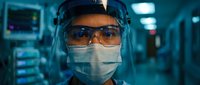 Female healthcare worker wearing protective equipment including face shield and surgical mask in modern hospital corridor with medical equipment background.