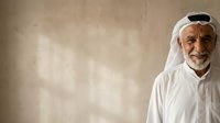 Senior Middle Eastern man wearing traditional white thobe and ghutra headwear smiles warmly against neutral beige background. Portrait suitable for business, healthcare, education, and cultural representation projects.