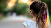 Young woman with rainbow colored hair streak in ponytail wearing white polka dot top, viewed from behind against blurred bokeh background in outdoor setting.