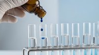 Laboratory technician in white gloves using dropper to add blue liquid sample into test tubes for scientific research and chemical analysis experiments.