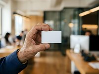 Senior male professional displaying blank business card in modern office environment with colleagues working in background.