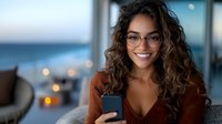 Young mixed race woman with curly hair and glasses smiling while using smartphone at home. Cozy evening atmosphere with bokeh lights and ocean view background.
