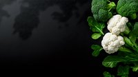 Fresh white cauliflower heads with vibrant green leaves against dark background, dramatic lighting creates contrast for food photography and healthy eating concept.