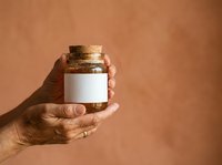 Person with medium skin tone holding small glass jar with cork stopper and blank white label against warm terracotta background for product mockup or cosmetic branding.