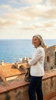 Mature Caucasian businesswoman enjoying peaceful moment on Mediterranean coastal terrace overlooking sea and historic architecture during golden hour.