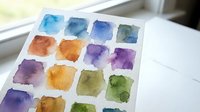 Watercolor paint palette with vibrant color swatches on white paper near window. Art supplies for creative painting and artistic expression projects.
